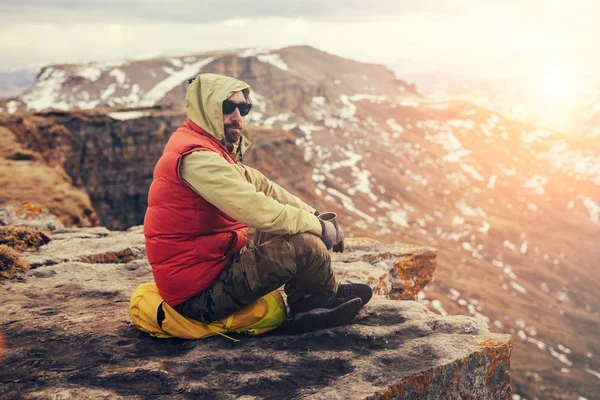 An adult bearded male traveler in a red jacket sits on the edge of a cliff, enjoying the mountain nature — Stock Photo, Image