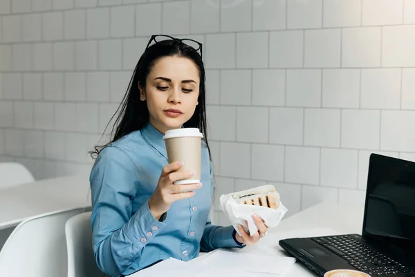 Zajęty dziewczyna student w niebieską koszulę, siedząc w kawiarni, Jedzenie Kanapki przed wykłady — Zdjęcie stockowe