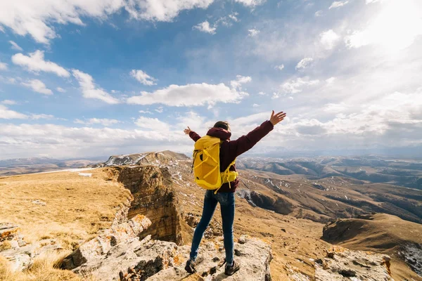 Menina ativa feliz viaja através das montanhas caucasianas com uma mochila amarela, levantou as mãos, goza — Fotografia de Stock