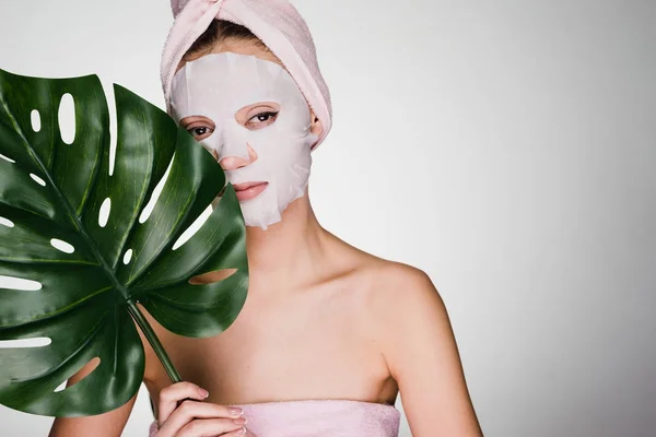 Κορίτσι με ένα ροζ πετσέτα στο κεφάλι της, στο πρόσωπό της έναν ιστό θρεπτική μάσκα, ημερήσιο σπα — Φωτογραφία Αρχείου
