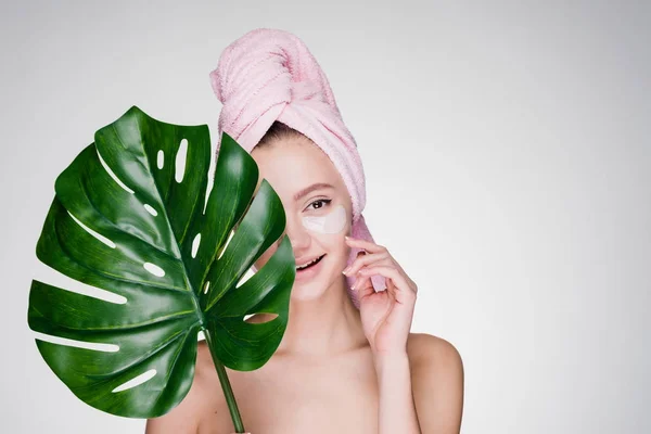 Joven feliz sonriente chica con toalla rosa en la cabeza sostiene una hoja verde, bajo los ojos parches — Foto de Stock