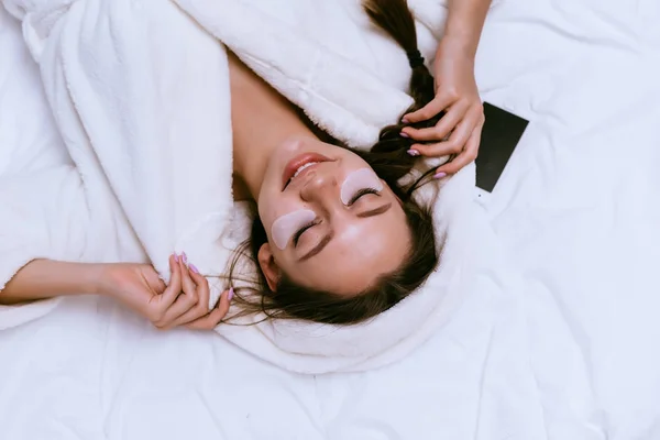 Jovem sorrindo menina encontra-se na cama, em jaleco branco, sob manchas olhos — Fotografia de Stock