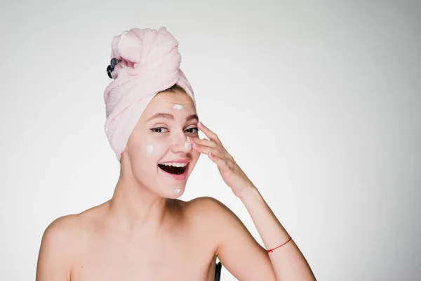 Χαριτωμένο νεαρή κοπέλα χαμογελαστή με πετσέτα στο κεφάλι, εφαρμογή ενυδατική κρέμα στο πρόσωπό — Φωτογραφία Αρχείου