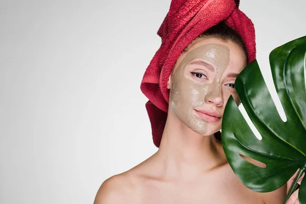 Een aantrekkelijk meisje met een rode handdoek op haar hoofd een klei masker toegepast op de helft van het gezicht, een spa-procedure — Stockfoto