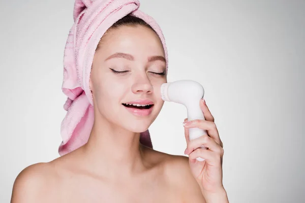 Young beautiful girl with a pink towel on her head makes a deep cleansing of the face skin with the help of an electric brush — Stock Photo, Image