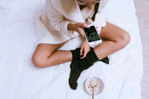 Uma menina em um vestido de vestir branco terry e meias quentes está sentado na cama e tirar fotos de seu café da manhã — Fotografia de Stock