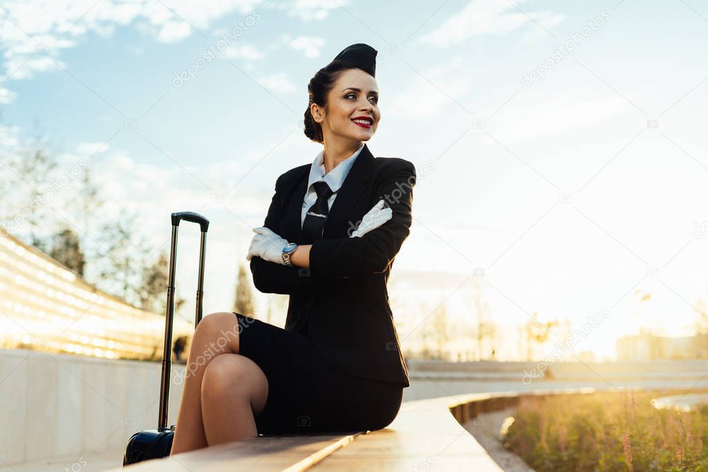 happy young stewardess girl in uniform sits in park, with suitcase, waiting for airplane