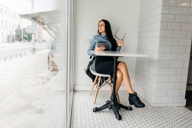 genç esmer kız mavi gömlekli kafede oturur, onun elinde bir tablet tutar ve pencereden görünüyor