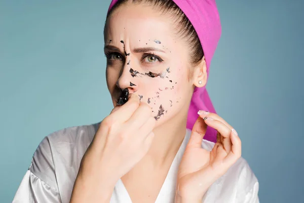 Una chica descontenta con una toalla rosa en la cabeza se quita una máscara de limpieza negra de la cara — Foto de Stock