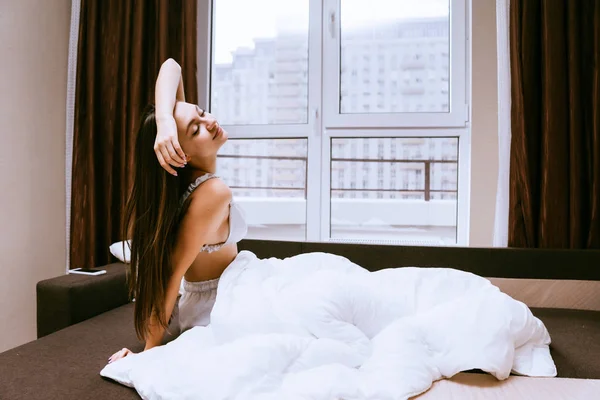 Uma jovem jaz na cama sob um cobertor branco, acordou de manhã cedo — Fotografia de Stock