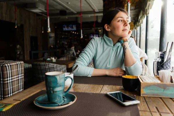 Una joven está sentada en un café, calentándose y mirando pensativamente por la ventana. — Foto de Stock