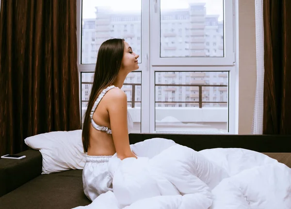 Bonito jovem menina sentado na cama sob um cobertor branco, não quer se levantar — Fotografia de Stock
