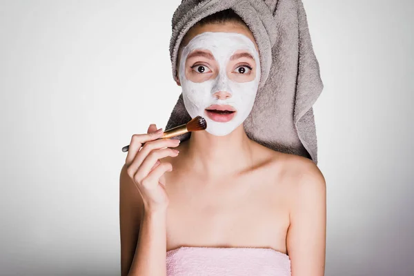 Encantadora chica sorprendida con una toalla en la cabeza se pone una máscara blanca nutritiva en la cara con la ayuda de un cepillo —  Fotos de Stock
