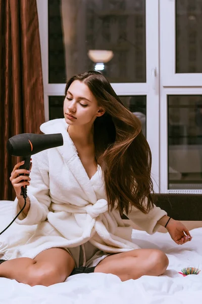 Красивая длинноволосая девушка в белом махровом халате сидит на кровати и сушит длинные волосы феном — стоковое фото