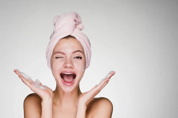 Happy joyful young girl with a pink towel on her head applies cleansing foam on her face — Stock Photo, Image