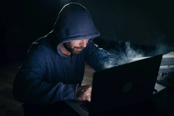 Um homem barbudo perigoso adulto em um capuz fazendo algo ilegal em um laptop, hackeando no escuro — Fotografia de Stock