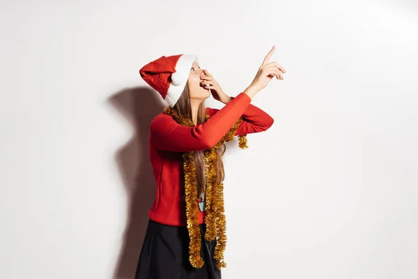 Una joven con una gorra roja como Santa Claus se sorprende al señalar con el dedo algo —  Fotos de Stock