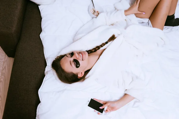 En ung jente i hvit morgenkåpe ligger på senga og ler under øynene på svarte flekker. – stockfoto