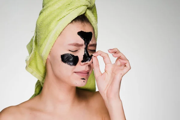 Молодая недовольная девушка с зеленым полотенцем на голове снимает черную очищающую маску с лица, ей неудобно — стоковое фото