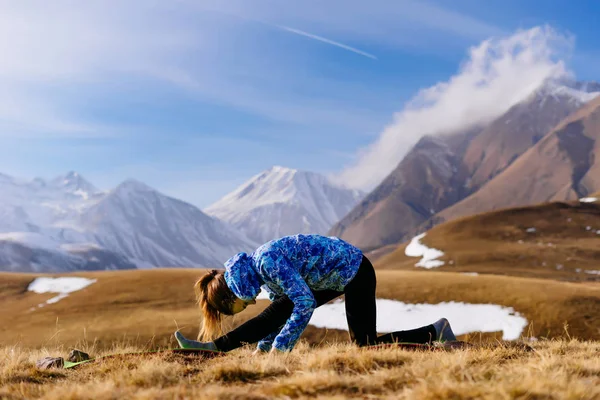 Активная молодая девушка в синей куртке занимается йогой на фоне гор Кавказа — стоковое фото