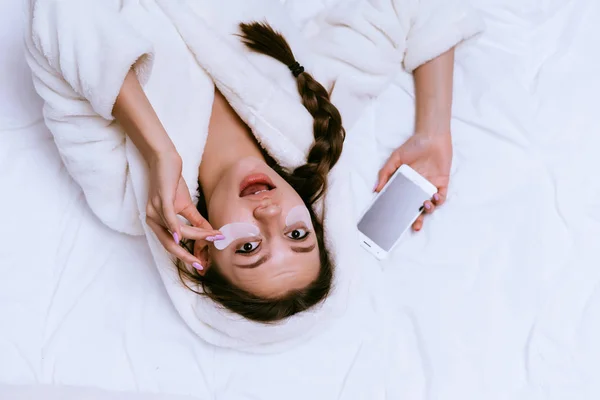 Удивленный молодой девушки лежит на кровати, держит телефон и снимает пластыри — стоковое фото