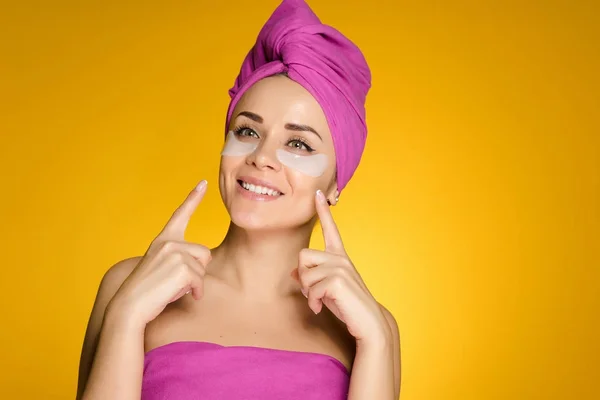 Felice ragazza sorridente con un asciugamano rosa sulla testa che punta le dita a toppe sotto gli occhi — Foto Stock