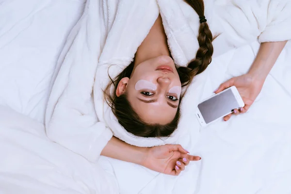 Piękna młoda dziewczyna w szlafrok biały frotte, leżąc w łóżku trzymając smartfon — Zdjęcie stockowe