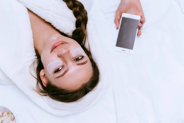 Niña con un vestido de terciopelo blanco está acostado en la cama, sosteniendo un teléfono inteligente — Foto de Stock