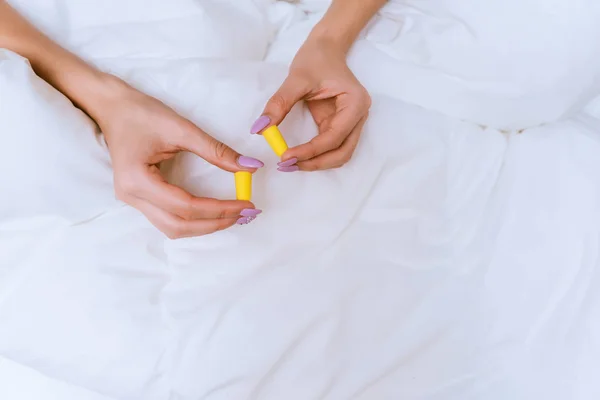 Het meisje zit op een bed onder een witte deken, gele oordoppen bezit tegen straatrumoer — Stockfoto