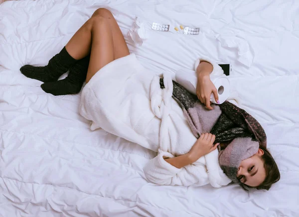 Грустная молодая девушка в белом махровом халате заболела из-за холодной погоды, лежит в постели, много таблеток — стоковое фото