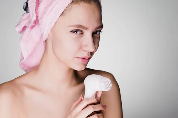 Funny mladá dívka s růžový ručník na hlavě drží Elektrický kartáč pro hloubkové čištění kůže na tváři — Stock fotografie