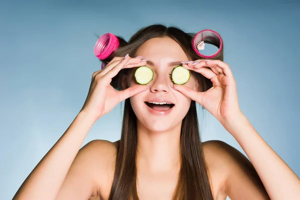 Happy krásná dívka drží kousky okurky pro zvlhčení pokožky v obličeji, na hlavě natáčky — Stock fotografie