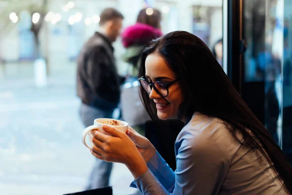 Смеющаяся милая девушка в очках сидит в кафе, наслаждается ароматным кофе — стоковое фото