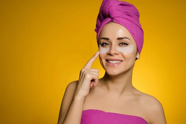 Όμορφη χαμογελαστή κοπέλα με ροζ πετσέτα στο κεφάλι της μοιάζει μετά το πρόσωπό της, κάτω από τα μάτια μπαλώματα — Φωτογραφία Αρχείου
