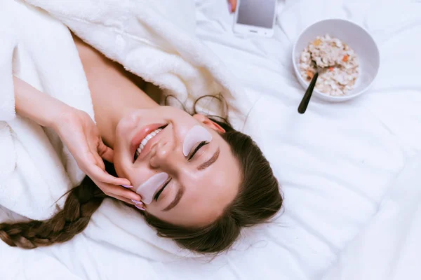 Cute girl uśmiechający się w białą szatę opiera się na łóżku z samego rana, tuż obok płyty Kasza płatki owsiane — Zdjęcie stockowe