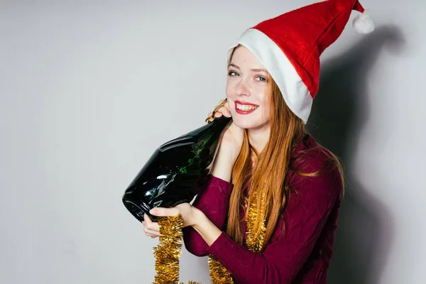 Ευτυχισμένος μεθυσμένος κοκκινομάλλα κοπέλα με κόκκινο καπάκι στο κεφάλι της, γιορτάζει το νέο έτος, κρατώντας ένα μπουκάλι σαμπάνια — Φωτογραφία Αρχείου