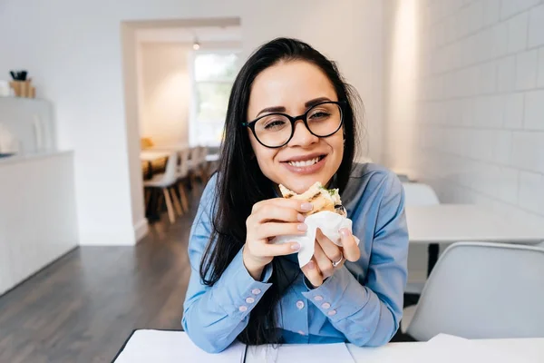 メガネと貪欲、カフェでサンドイッチを食べる青いシャツを着て幸せ空腹女学生 — ストック写真