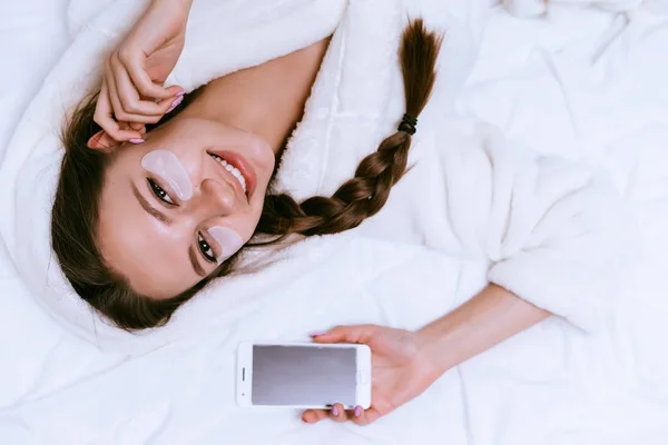 Счастливая милая девушка лежит в постели, рано утром, держа смартфон — стоковое фото