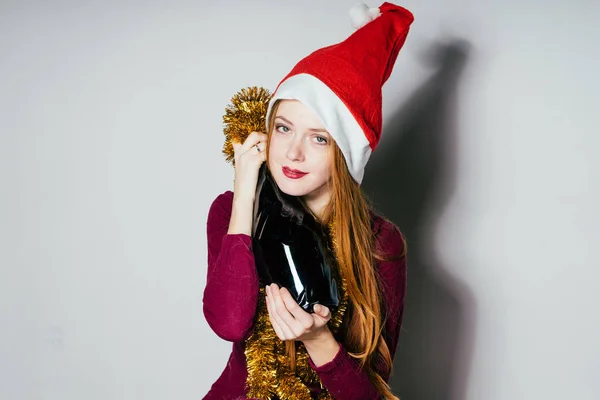 Douce fille aux cheveux roux dans un bouchon rouge comme le Père Noël étreint une bouteille de champagne, atmosphère du Nouvel An — Photo