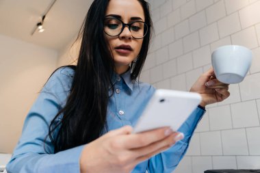meşgul genç kız mavi gömlek ve gözlük kahve içme ve onun smartphone arıyorum
