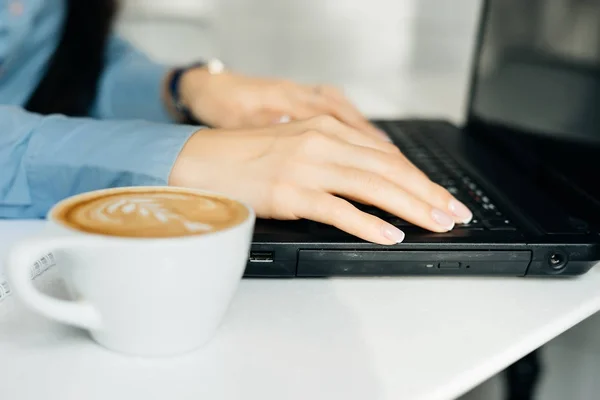 Девушка фрилансер в синей рубашке работает на ноутбуке, далее кружка с горячим кофе — стоковое фото
