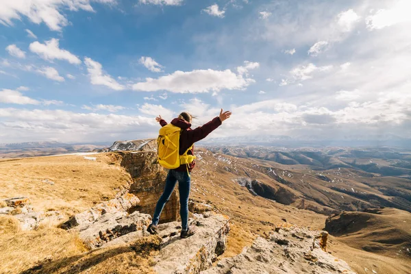 Happy active girl travels through the Caucasian mountains with a yellow backpack, handles raised upwards — Stock Photo, Image