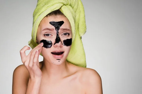 En ung flicka med en grön handduk på huvudet gäller en svart cleansing mask på problemområden i ansiktet — Stockfoto