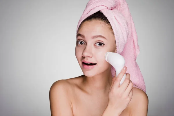 Divertente ragazza carina con un asciugamano rosa sulla testa facendo una profonda pulizia della pelle del viso spazzola elettrica — Foto Stock