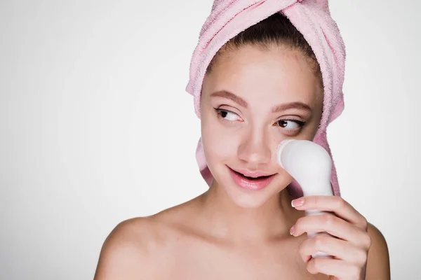 Roztomilá mladá dívka s růžový ručník na hlavu dělat hloubkové čištění kůže obličeje s elektrickým kartáčem — Stock fotografie