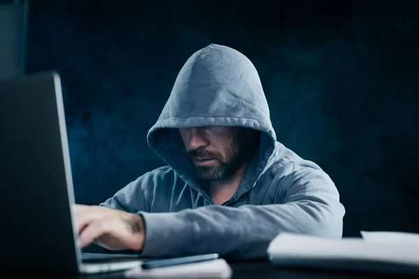 Um jovem hacker senta-se atrás de um laptop sob um capuz — Fotografia de Stock