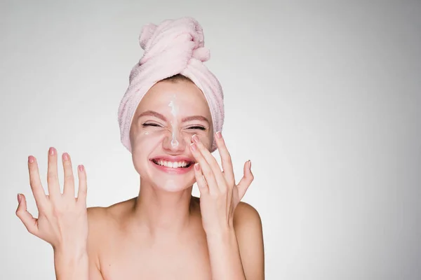 Laughing young girl with a pink towel on her head applying a nourishing cream on her face — Stock Photo, Image