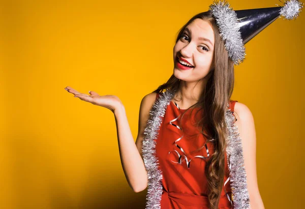 Een schattige lachende meisje in een rode jurk en met een zilver klatergoud rond haar nek vieren het nieuwe jaar, iets met de hand tonen — Stockfoto