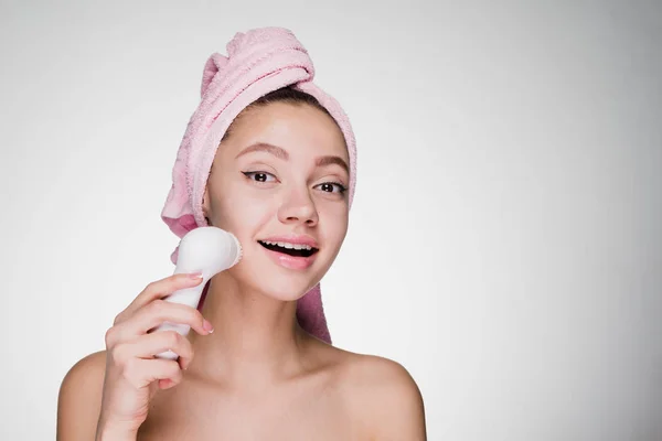 Счастливая улыбающаяся женщина с розовым полотенцем на голове очищает лицо электрической щеткой — стоковое фото