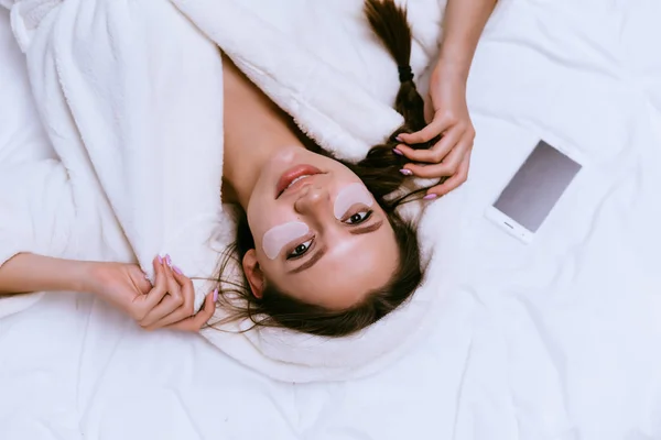 Młoda dziewczyna w szlafrok biały frotte leży na łóżku, pod oczy patch — Zdjęcie stockowe